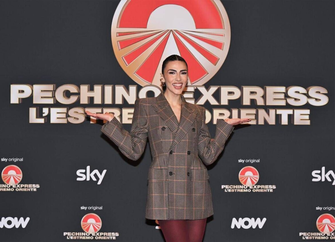 Woman posing on a red-carpet backdrop with arms raised, wearing a plaid blazer and burgundy tights; event logos read 'Pechino Express' and 'Sky' in the background.